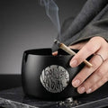 Black ashtray with artistic design, featuring a hand holding a lit cigarette.