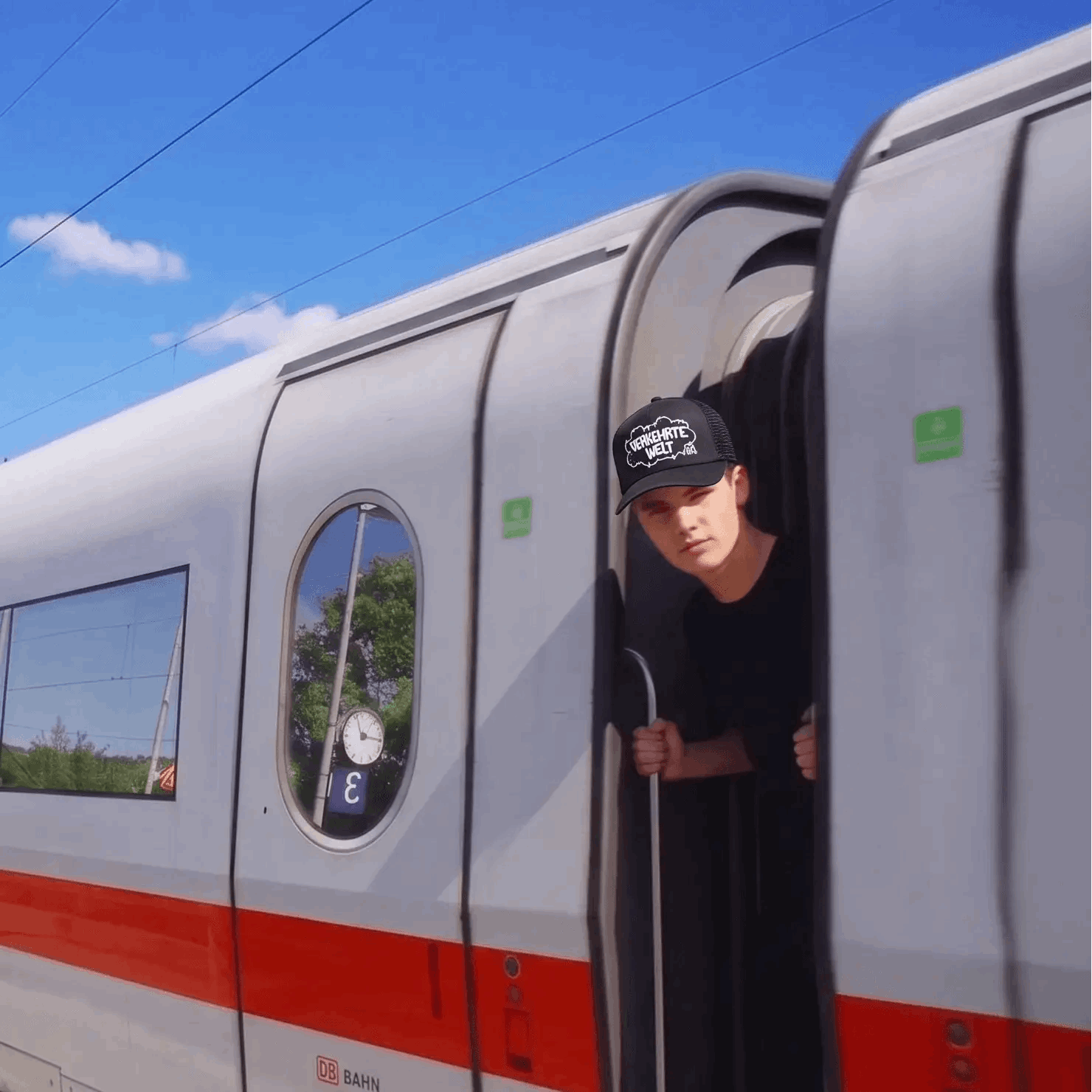 Train with a red stripe featuring a person leaning out of the door, under a clear blue sky.