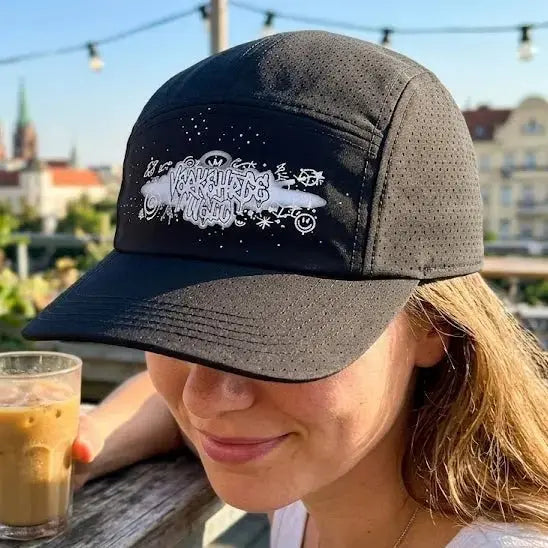 Black cap with graphic design featuring playful text and illustrations, worn by a woman outdoors.